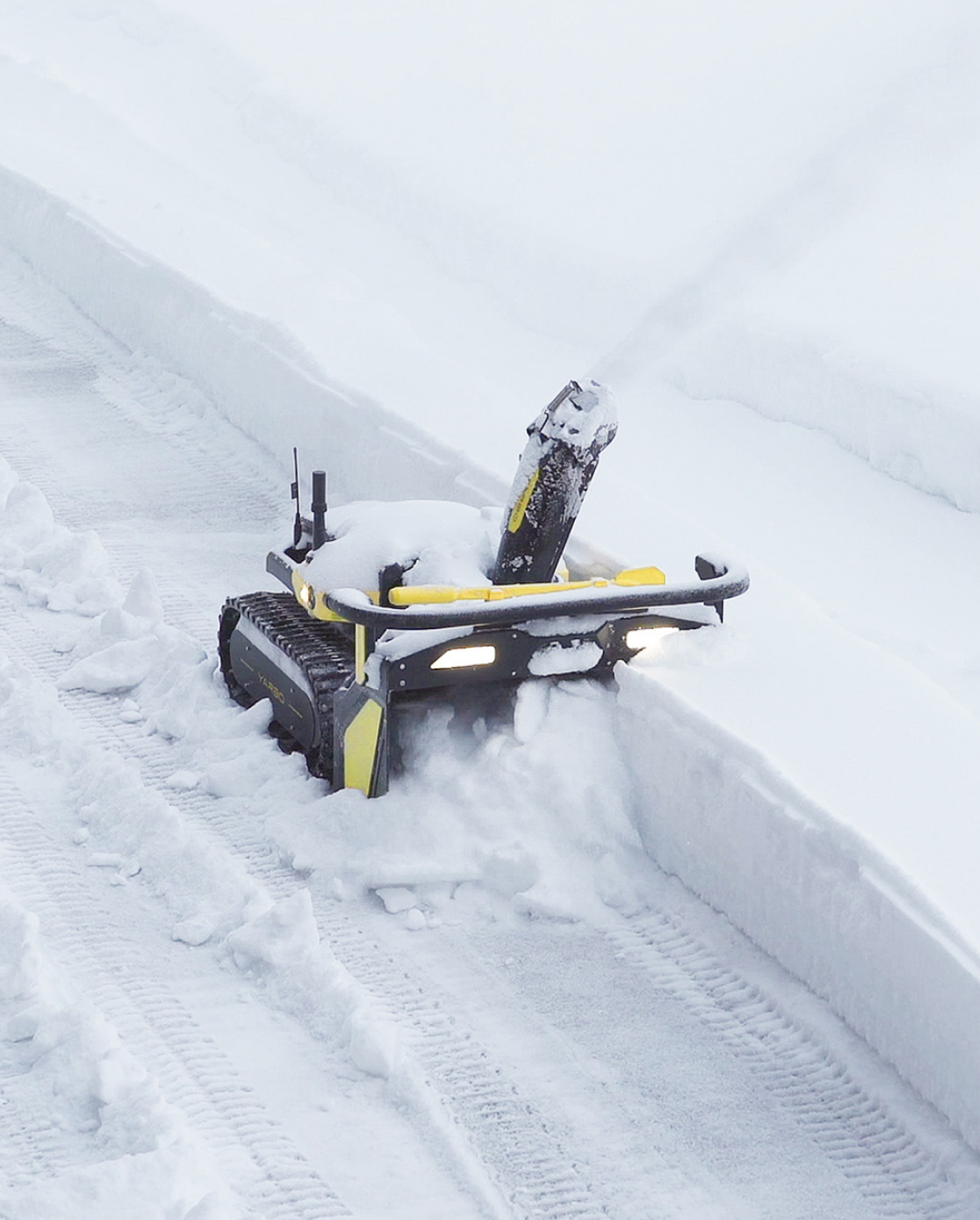 Black and yellow robotic snow blower by Yarbo clearing snow, tracked base, front chute, illuminated lights, side view in snowy driveway